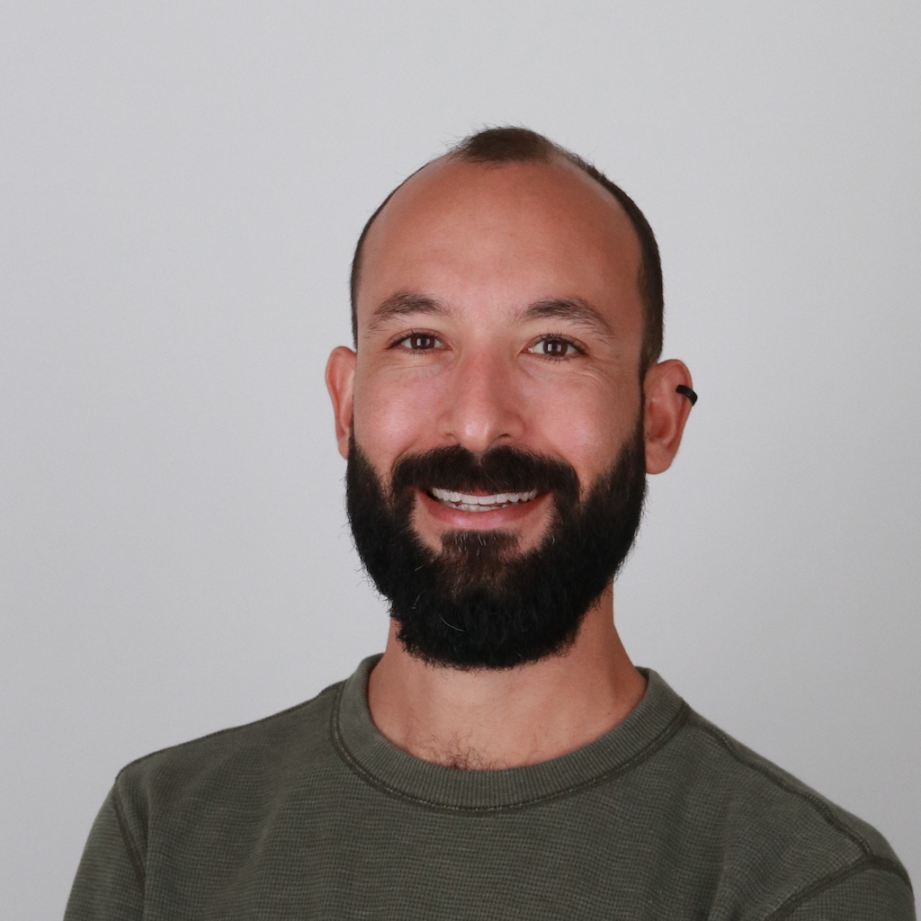 A bearded man in a green shirt smiling in front of a white background.